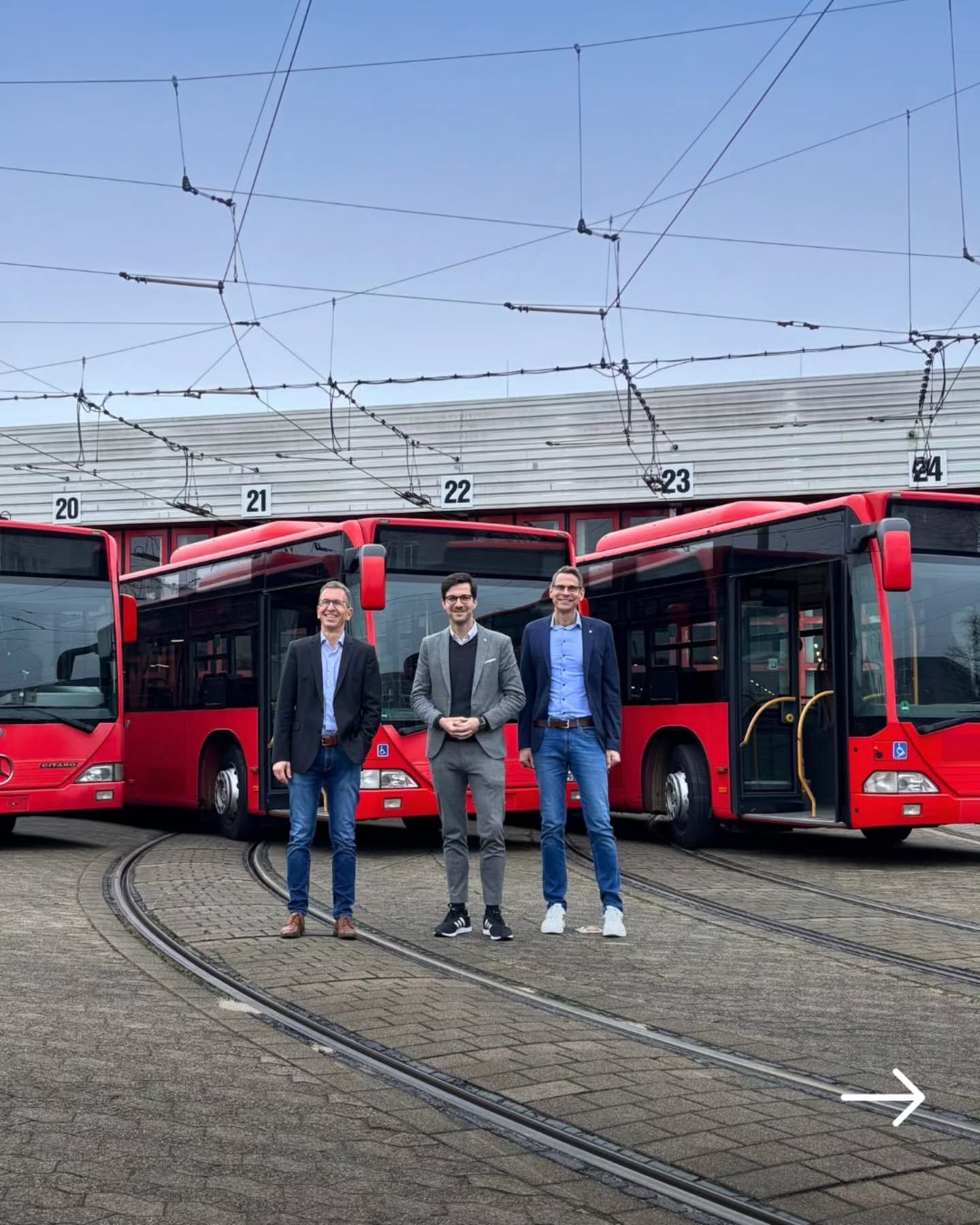Dieser Alternativtext wurde KI-generiert: Drei Personen stehen vor roten Bussen in einem Busdepot. Im Hintergrund sind weitere Busse und Stromleitungen zu sehen.