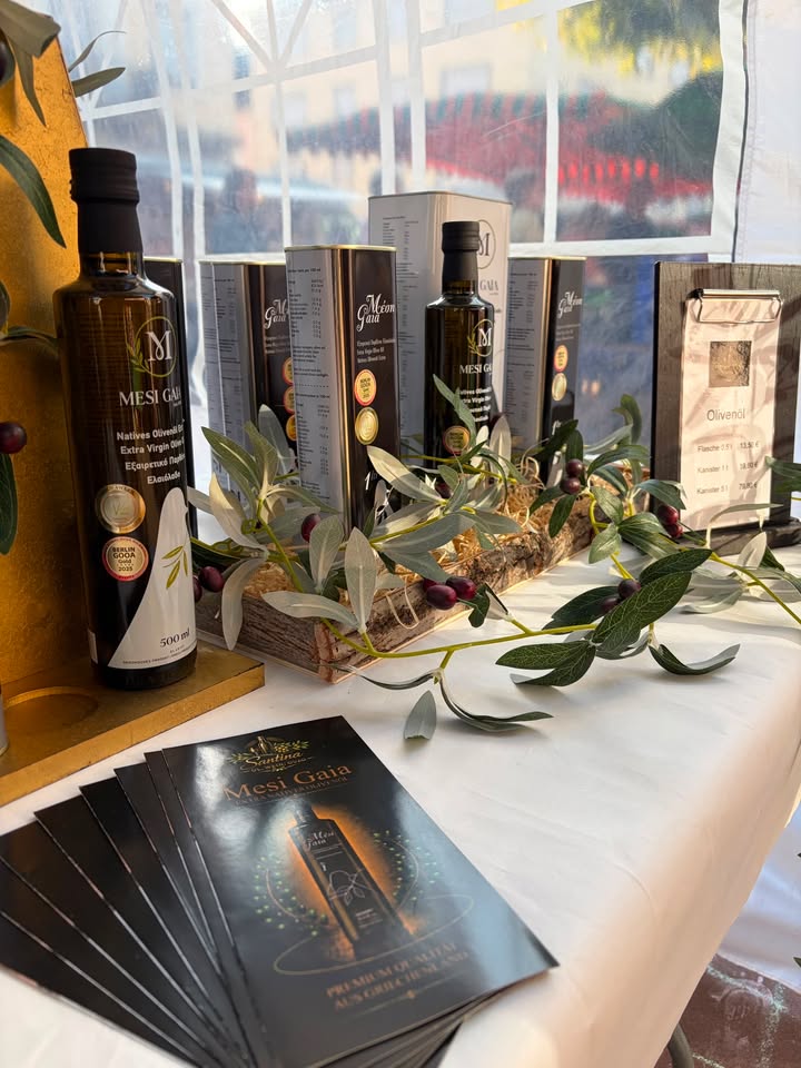Ein Tisch mit Flaschen und Kartons der Marke Mesi Gaia, die Natives Olivenöl Extra Virgin enthalten. Davor liegen Broschüren mit der Aufschrift Premium Qualität aus Griechenland. Olivenzweige und eine Preistafel für Olivenci vervollständigen die Präsentation. (KI-Alternativtext)