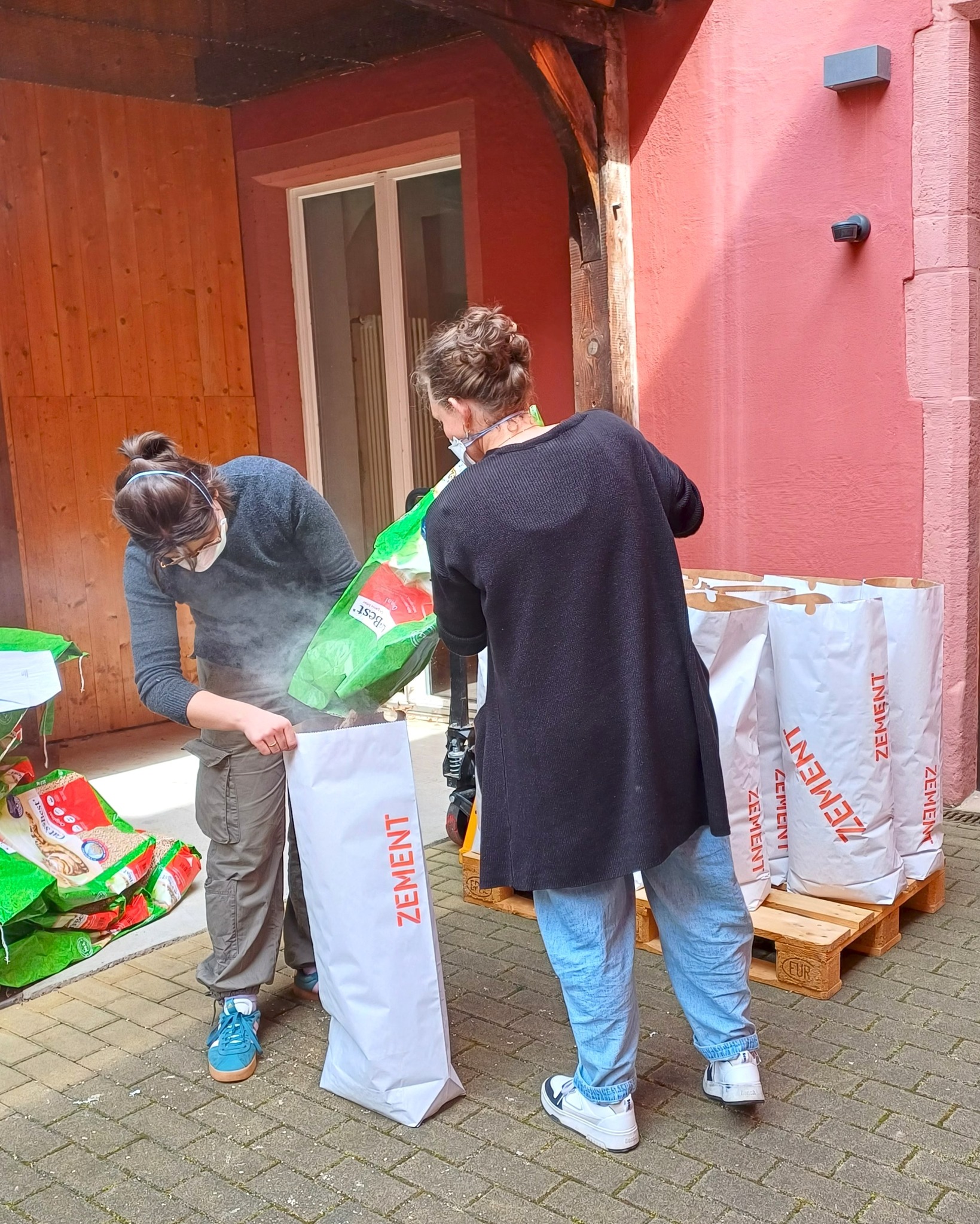 Zwei Personen füllen einen weißen Sack mit der Aufschrift „ZEMENT“ aus einem grünen Beutel, während Staub aufsteigt. Im Hintergrund stehen weitere Zementsäcke auf einer Palette vor einer roten Wand. (KI-Alternativtext)