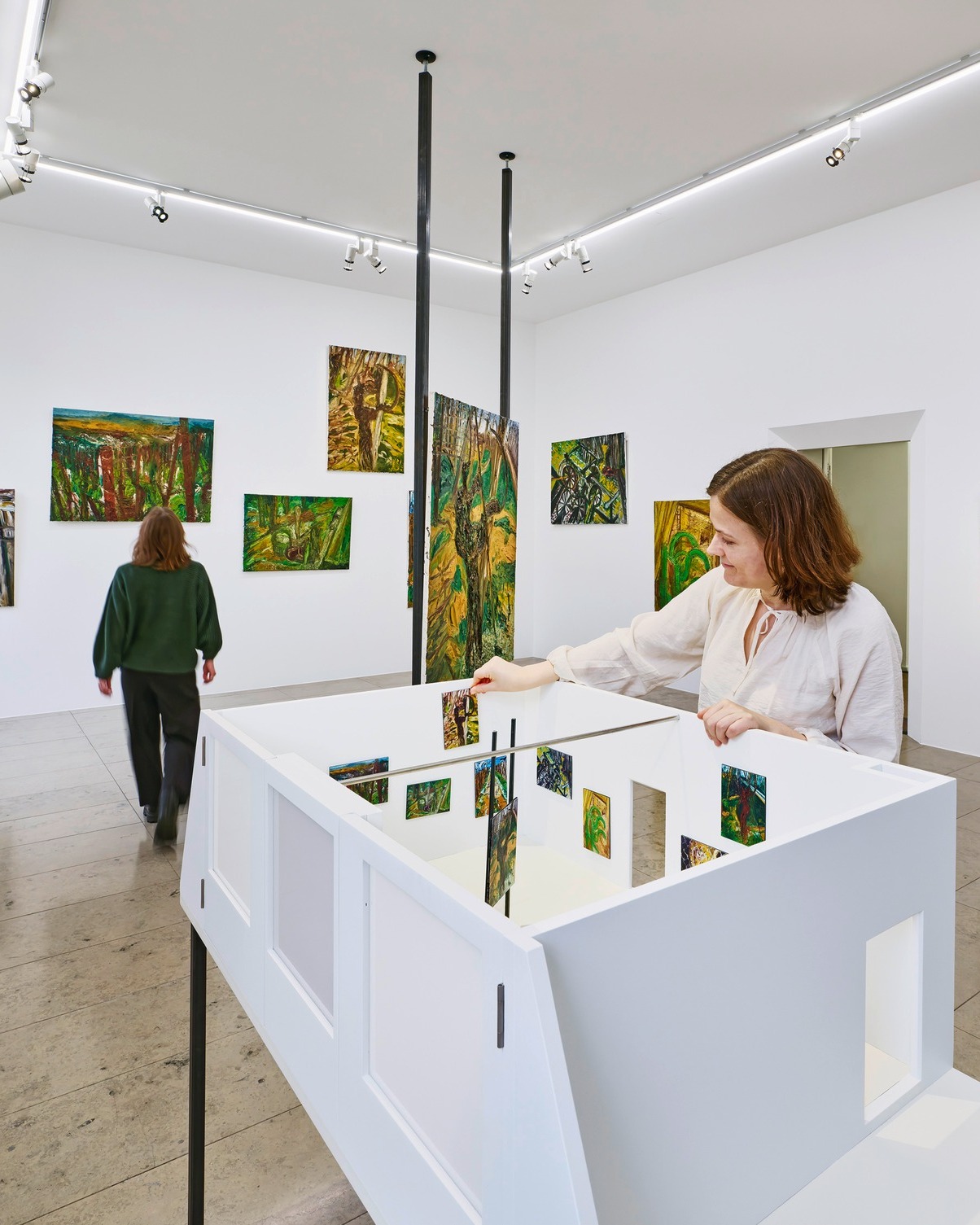 Dieser Alternativtext wurde KI-generiert: Ein heller Raum mit Kunstwerken an den Wänden und einer Person, die sich eine Installation betrachtet. Eine weitere Person steht dem Rücken zugewandt und betrachtet ebenfalls Kunstwerke.