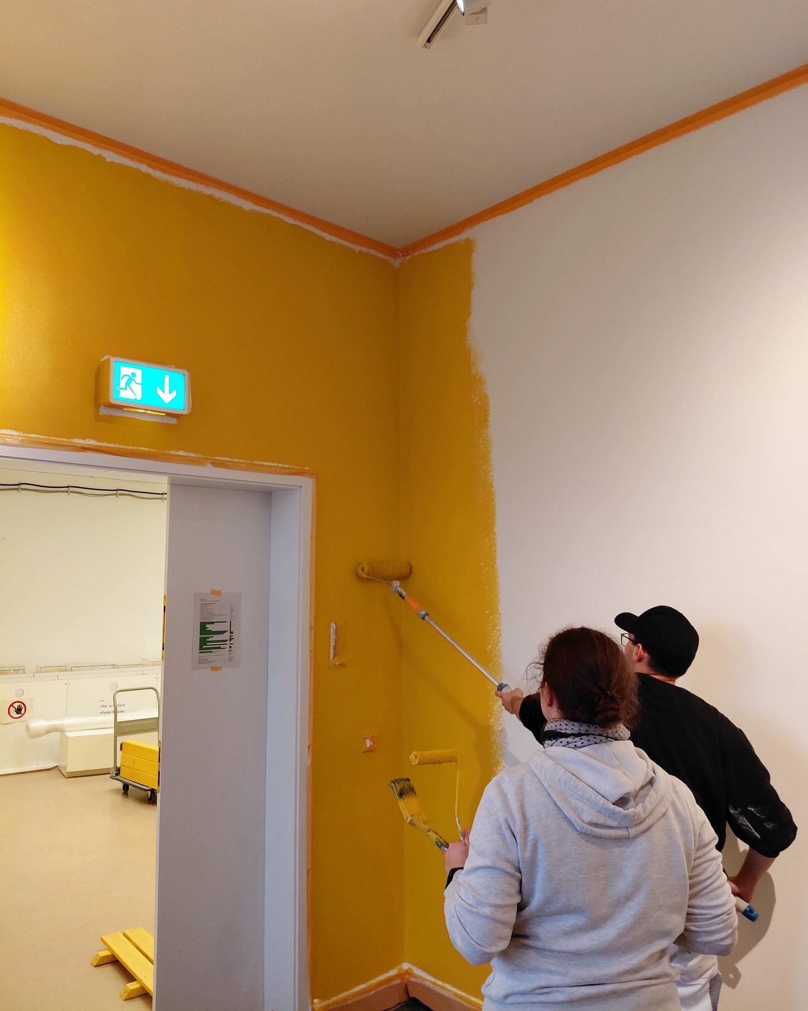 Dieser Alternativtext wurde KI-generiert: Eine Wand wird gestrichen. Ein Türrahmen und ein Notausgangsschild sind ebenfalls im Bild zu sehen.