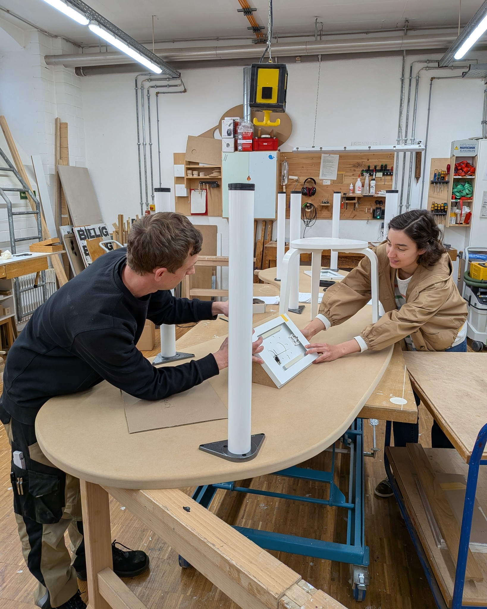 Dieser Alternativtext wurde KI-generiert: Zwei Personen arbeiten in einem Werkraum an einem Tischbauprojekt. Im Hintergrund sind Werkzeuge und Materialien an den Wänden zu sehen.