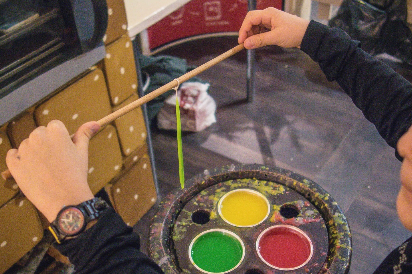 Eine Person hält einen Stock, an dem ein farbiges Wachsstück befestigt ist, über mehreren kleinen Behältern mit buntem Wachs.