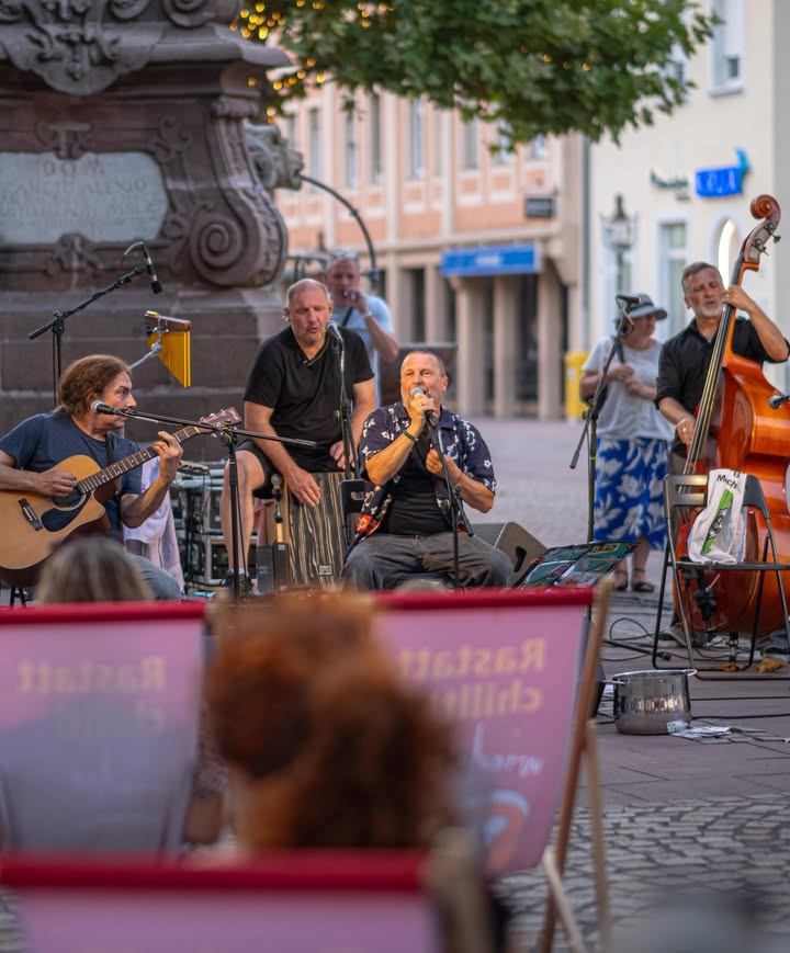 Mehrere Musiker spielen auf einer gepflasterten Straße vor einem Steinmonument. Ein Musiker spielt Akustikgitarre, ein anderer singt in ein Mikrofon und ein dritter spielt Kontrabass. Im unscharfen Vordergrund steht ein rosa Schild mit der Aufschrift "Rastatt Chillout". (KI-Alternativtext)