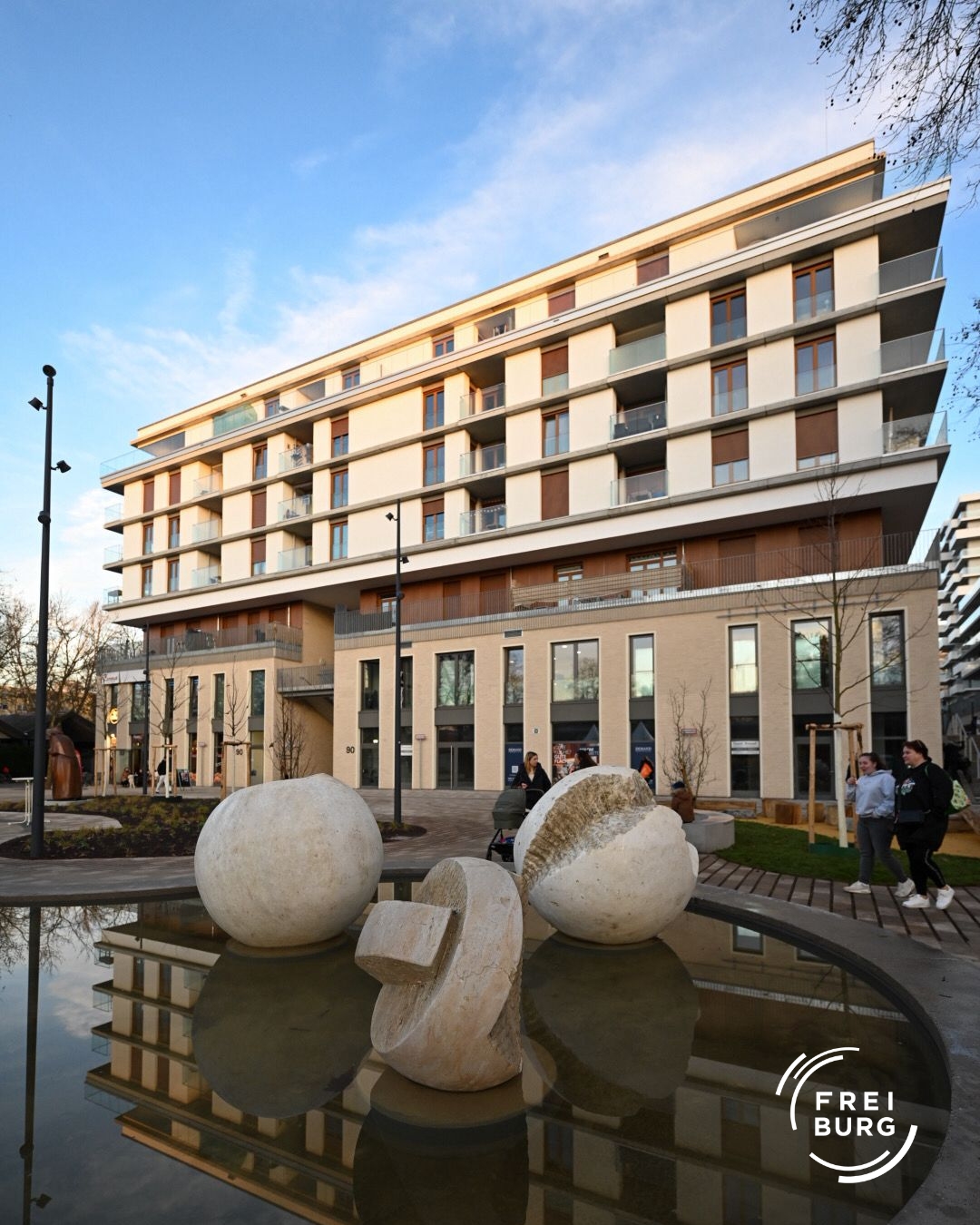 Dieser Alternativtext wurde KI-generiert: Ein modernes Gebäude mit vielen Fenstern steht im Vordergrund. Im unteren Bildbereich befinden sich Skulpturen in einem Wasserbecken.