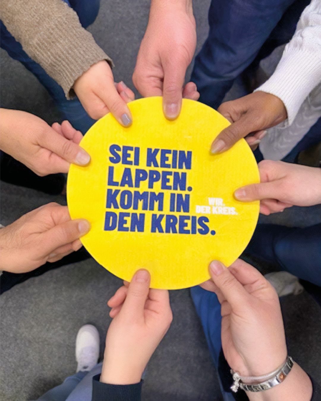 Dieser Alternativtext wurde KI-generiert: Mehrere Hände halten ein gelbes Schild mit blauem Text. Das Schild enthält eine Botschaft, die zur Teilnahme an einer Gemeinschaft einlädt.