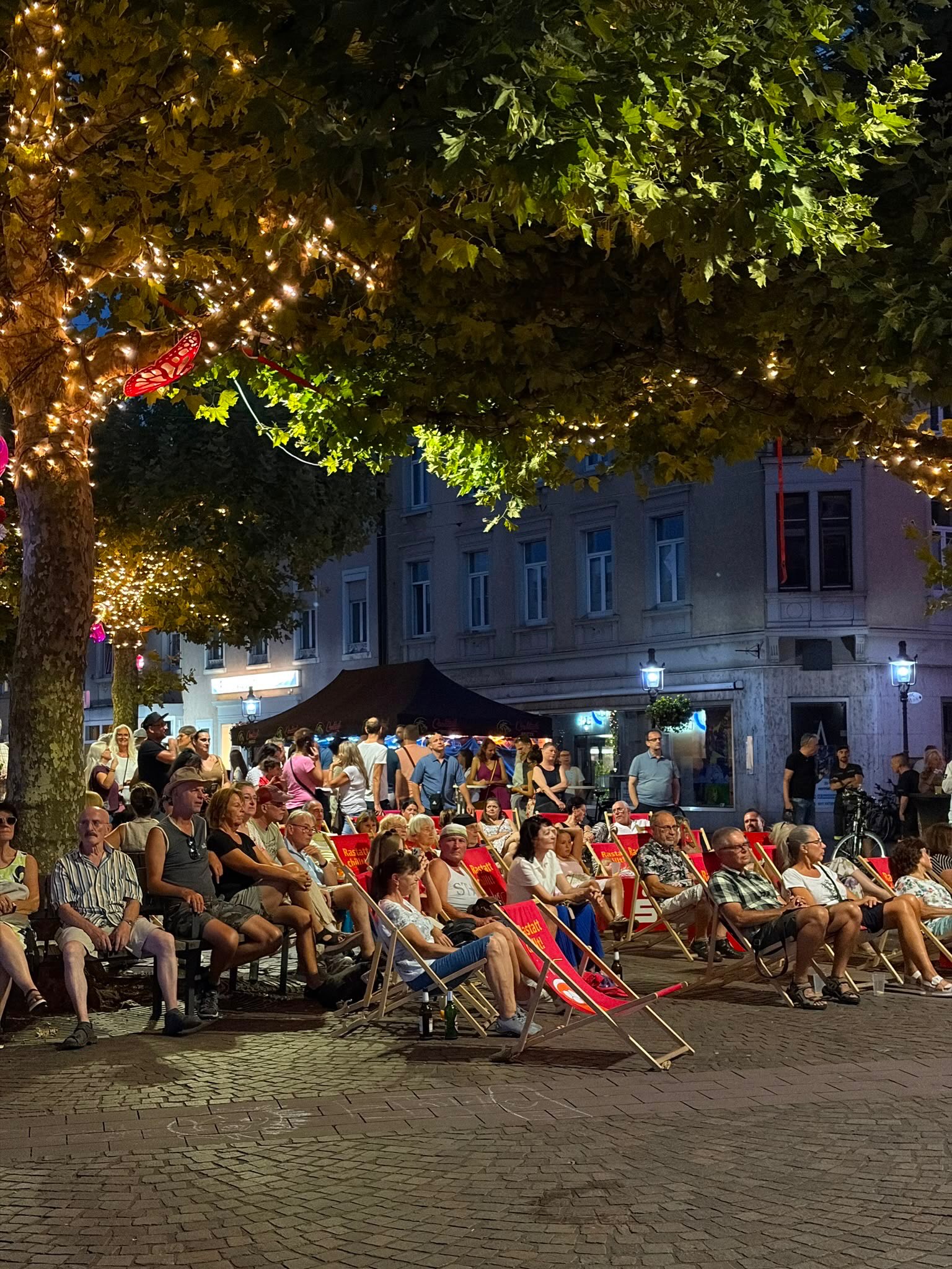Menschen sitzen nachts auf einem gepflasterten Platz in Reihen von roten Liegestühlen und auf Bänken, während sie in eine Richtung blicken. Große Bäume mit warmen Lichterketten rahmen die Szene ein, im Hintergrund stehen beleuchtete Gebäude und ein schwarzes Zelt. Auf den Liegestühlen ist der Schriftzug „Rastatt China“ zu erkennen. (KI-Alternativtext)