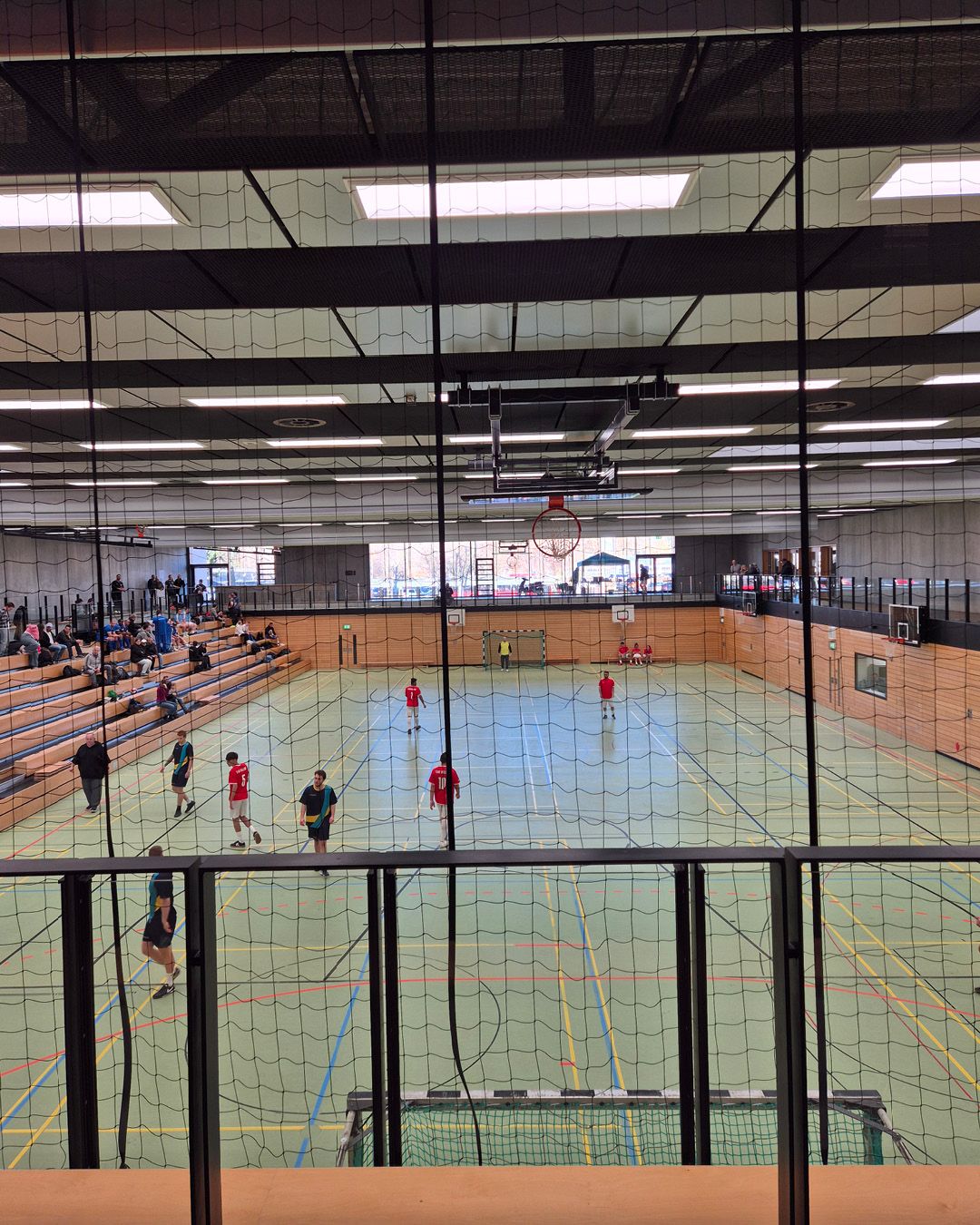 Dieser Alternativtext wurde KI-generiert: Ein helles Sportgebäude mit einer Holzbodenfläche und mehreren Personen, die Sport treiben. Durch ein Netz im Vordergrund ist der Blick auf die Halle möglich.