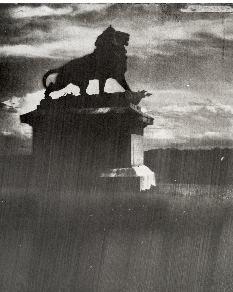Schwarze Silhouette einer Löwenstatue auf einem Sockel vor bewölktem Himmel, im Vordergrund Regenstriche. (KI-Alternativtext)