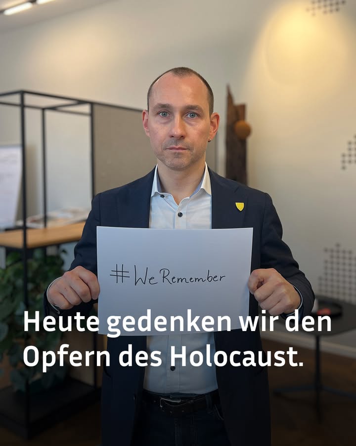 Dieser Alternativtext wurde KI-generiert: Eine Person hält ein weißes Schild mit einem Hashtag und Text in der Hand. Im Hintergrund sind Büroeinrichtungen zu sehen.