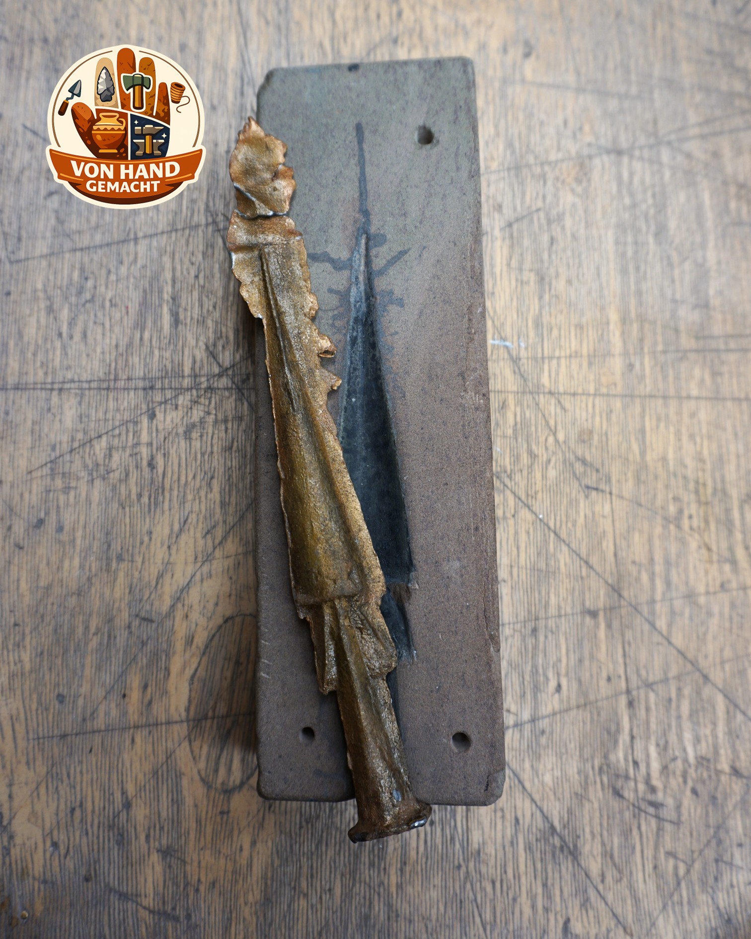 Ein rechteckiger Metallgussblock mit einer länglichen Aussparung liegt auf einer hölzernen Arbeitsfläche. Daneben liegt ein gegossenes Objekt in Form eines Schwertes mit rauer, bronzefarbener Oberfläche. Oben links ist ein rundes Logo mit dem Schriftzug "VON HAND GEMACHT" zu sehen. (KI-Alternativtext)