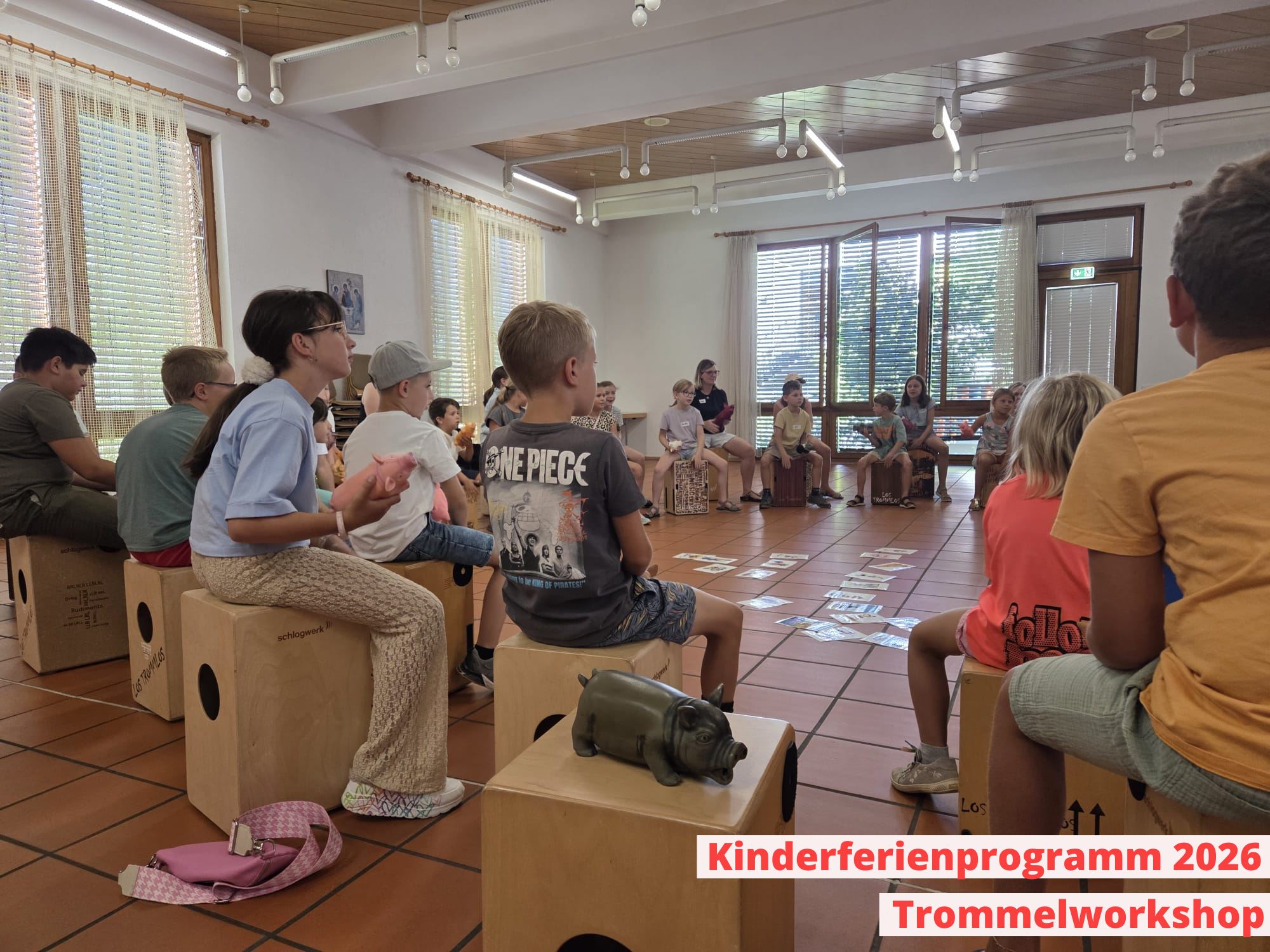 Eine Gruppe von Kindern sitzt im Kreis auf hölzernen Boxen in einem hellen Raum. Im Vordergrund steht eine kleine Tierfigur auf einer Box. Ein roter Text unten rechts lautet "Kinderferienprogramm 2026 Trommelworkshop". (KI-Alternativtext)