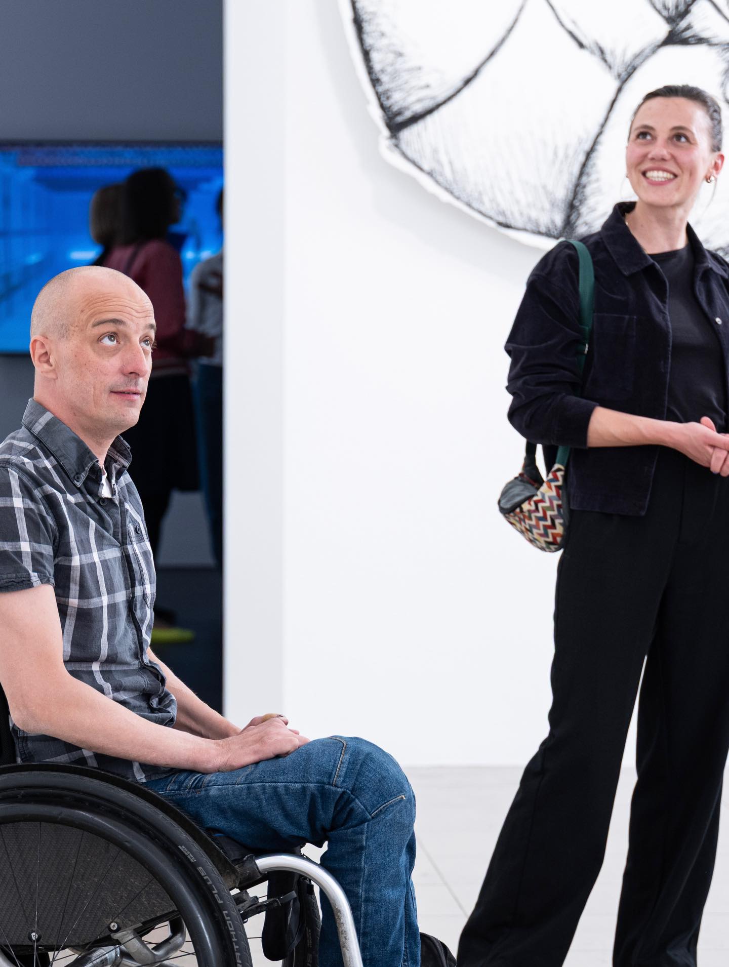 Dieser Alternativtext wurde KI-generiert: Zwei Personen befinden sich in einer hellen Galerie. Eine Person sitzt im Rollstuhl und blickt schauend zur zweiten Person, die steht und lächelt.