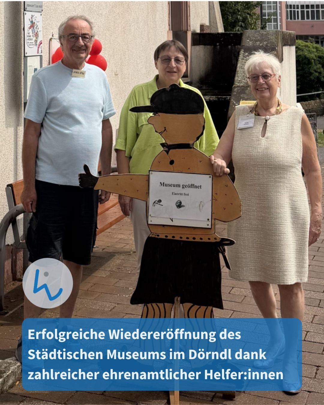 Drei Personen stehen im Freien vor einem Gebäude und flankieren eine hölzerne Figur mit dem Schild „Museum geöffnet, Eintritt frei“. Ein blauer Textbalken unten lautet: „Erfolgreiche Wiedereröffnung des Städtischen Museums im Dörndl dank zahlreicher ehrenamtlicher Helfer:innen“. (KI-Alternativtext)