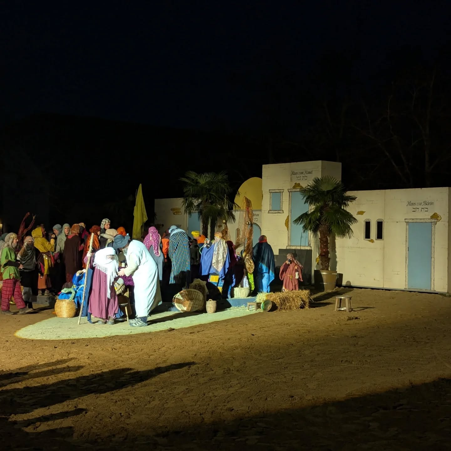 Dieser Alternativtext wurde KI-generiert: Eine Szene in der Nacht zeigt eine Gruppe von Menschen vor einem Gebäude mit architektonischen Details und Palmen. Das Bild ist in dunklen Tönen gehalten, mit Lichtquellen, die bestimmte Bereiche beleuchten.