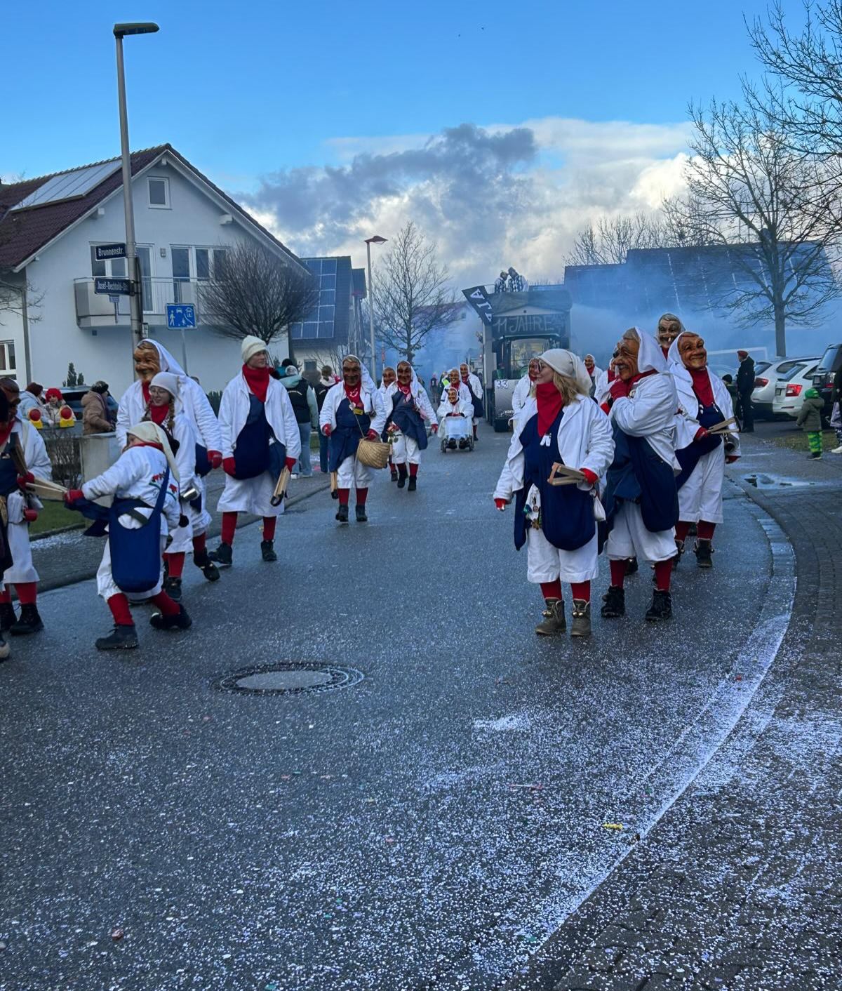 Dieser Alternativtext wurde KI-generiert: Eine Gruppe von Personen in weißen und roten Kostümen marschiert auf einer Straße. Im Hintergrund sind Gebäude und Bäume unter einem blauen Himmel zu sehen.