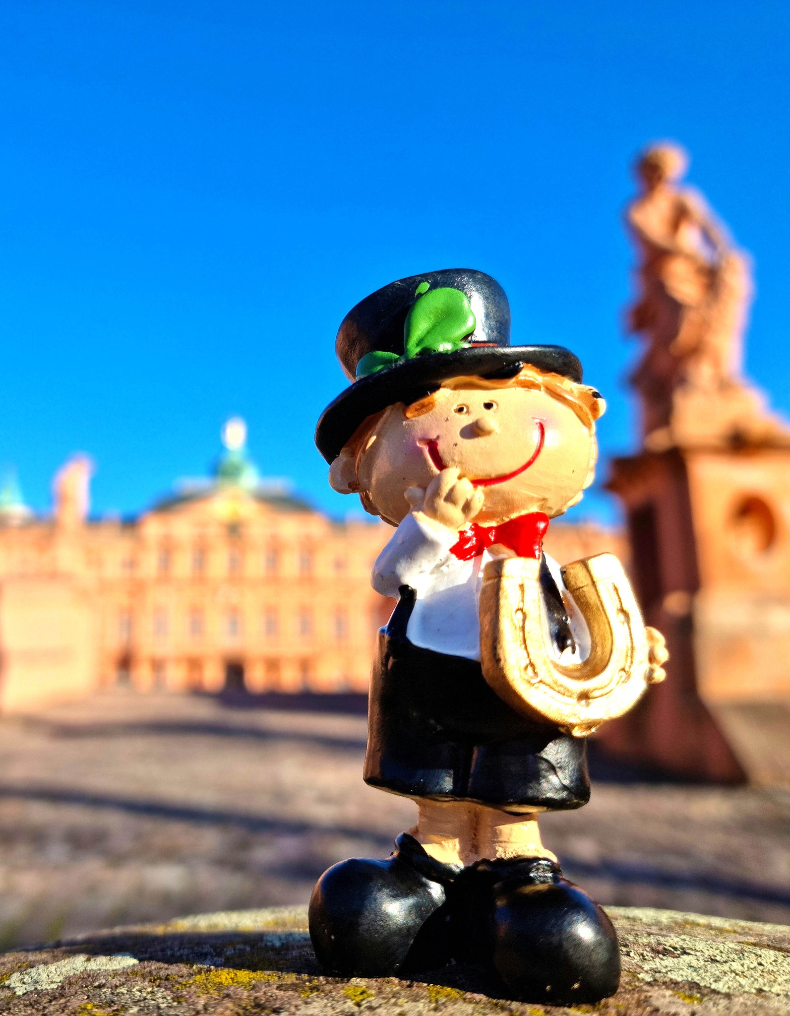 Dieser Alternativtext wurde KI-generiert: Eine kleine Figur mit Hut und Flöte steht im Vordergrund. Im Hintergrund ist eine Stadtlandschaft unter blauem Himmel zu erkennen.