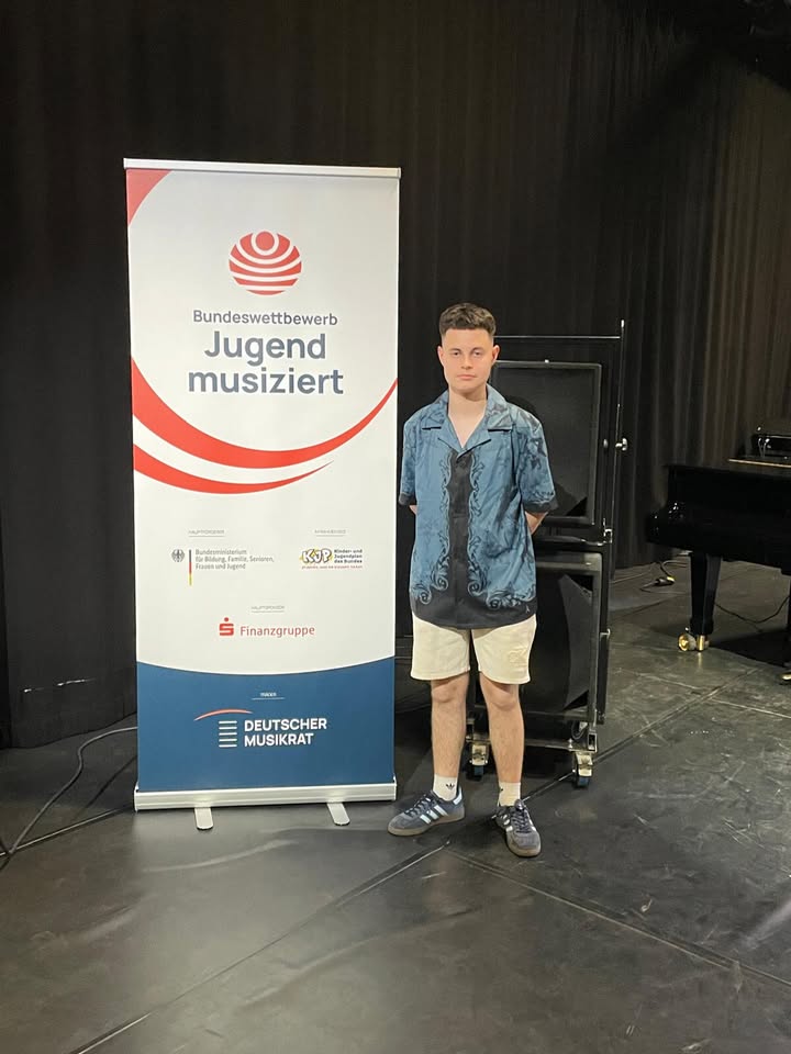 Eine Person steht vor einem Roll-Up-Banner mit der Aufschrift „Bundeswettbewerb Jugend musiziert“ und Logos von Förderern wie dem Deutschen Musikrat und der Sparkassen-Finanzgruppe. Im Hintergrund ist ein Flügel und dunkle Bühnengardinen zu sehen. (KI-Alternativtext)