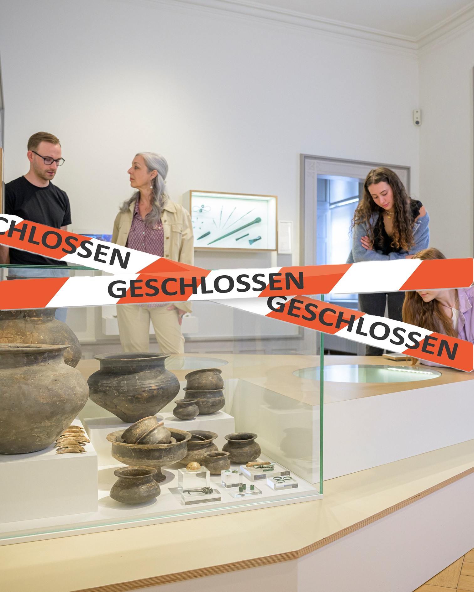 Ein Glasvitrinen mit antiken Tongefäßen und kleinen Artefakten steht im Vordergrund. Ein rot-weißes Absperrband mit der Aufschrift "GESCHLOSSEN" verläuft quer über die Vitrine. Im Hintergrund betrachten vier Personen die Ausstellung. (KI-Alternativtext)