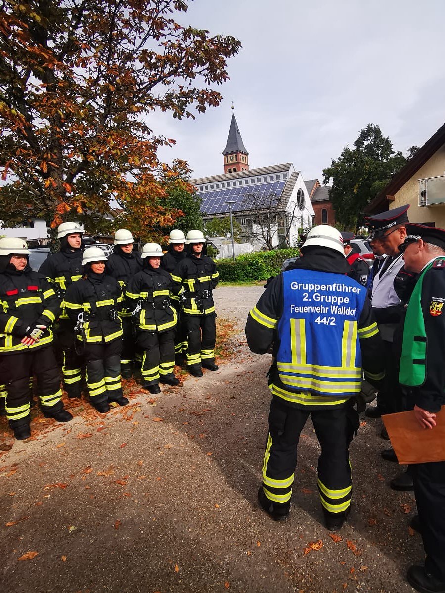 Dieser Alternativtext wurde KI-generiert: Eine Gruppe von Personen in Uniformen steht im Freien zusammen. Im Hintergrund ist ein Gebäude mit einem Kirchturm zu sehen.