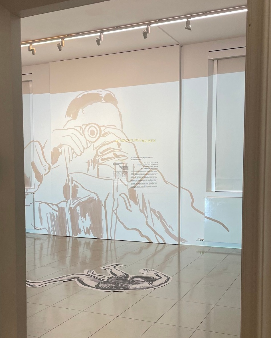 Dieser Alternativtext wurde KI-generiert: Ein heller Raum mit einer weißen Wand, auf der eine grafische Darstellung zu sehen ist. Text und Zeichnungen sind in hellem Braunton auf der Wand angebracht.