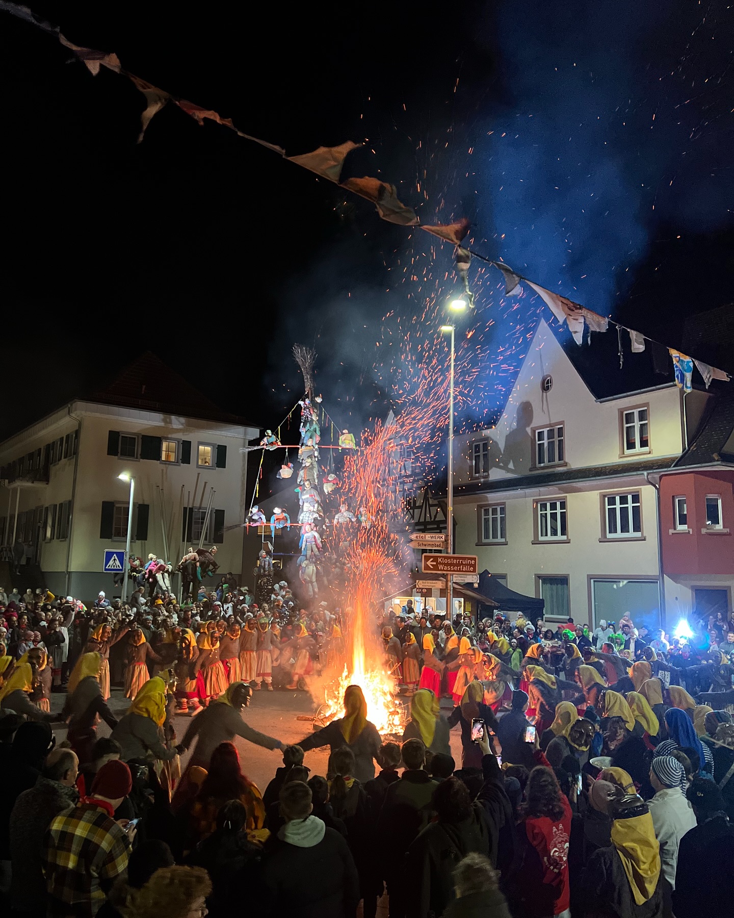 Das Foto zeigt eine Menschenmenge, die sich um ein Lagerfeuer versammelt hat, wobei im Hintergrund Gebäude und Dekorationen zu sehen sind. Rauchsäulen und brennende Partikel steigen in den Nachthimmel auf.