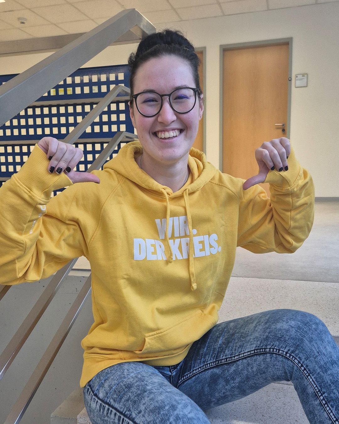 Dieser Alternativtext wurde KI-generiert: Eine Person sitzt auf einer Treppe und lächelt in die Kamera. Sie trägt einen gelben Kapuzenpullover mit Aufschrift und eine Brille.