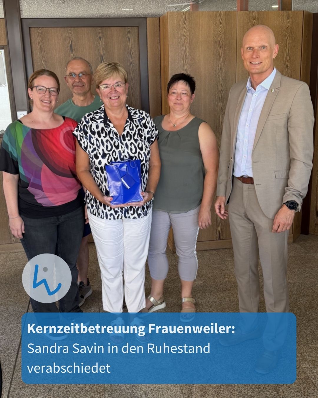 Fünf Personen stehen nebeneinander vor einer Holztür, wobei die Person in der Mitte eine blaue Geschenktüte hält. Ein Textfeld am unteren Rand lautet: „Kernzeitbetreuung Frauenweiler: Sandra Savin in den Ruhestand verabschiedet“. (KI-Alternativtext)