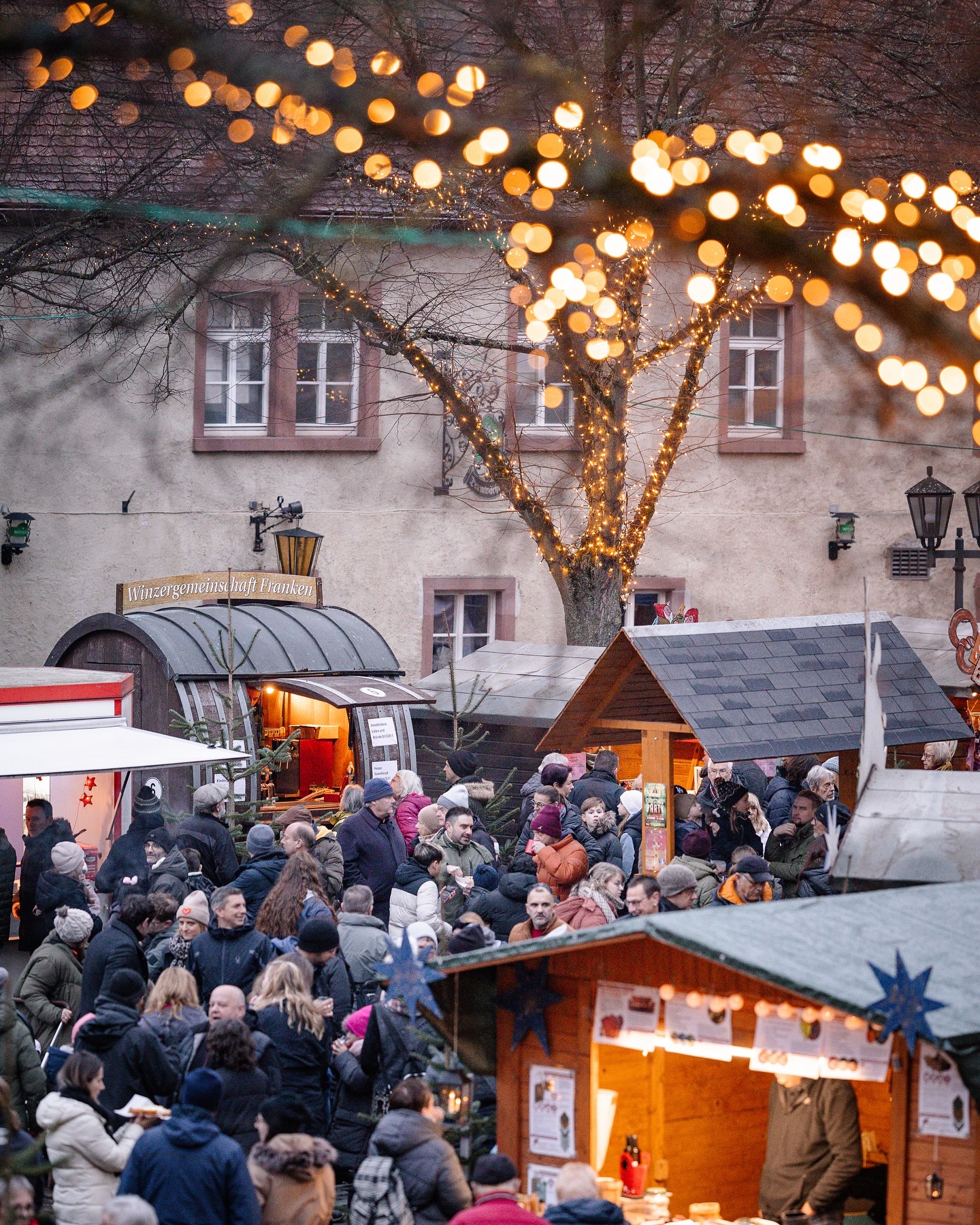 Ein Weihnachtsmarkt mit vielen Buden und Menschen. Lichterketten schmücken die Bäume.