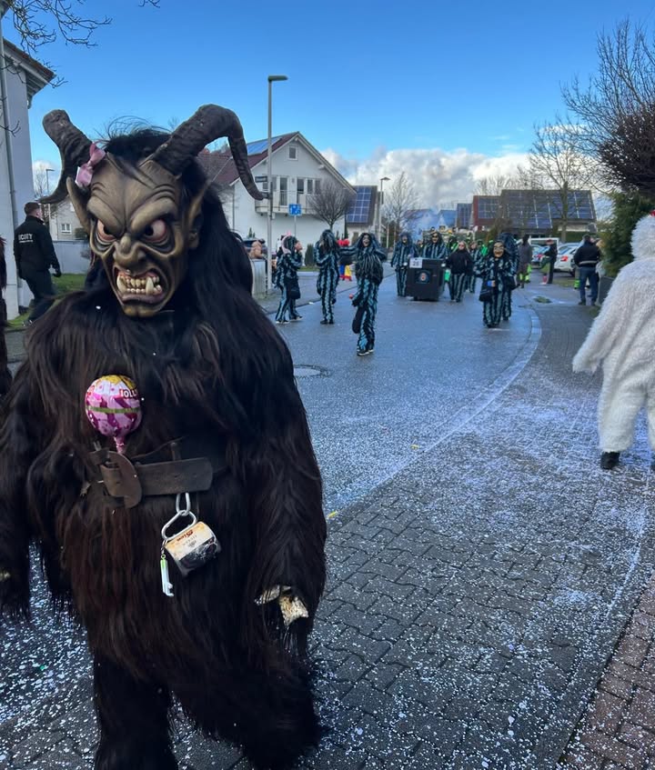 Dieser Alternativtext wurde KI-generiert: Eine Gruppe von Personen in auffälligen, dunklen Kostümen mit Masken steht und geht auf einer Straße. Im Hintergrund sind weitere Figuren und Gebäude zu sehen.