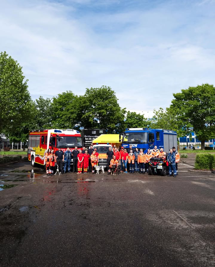 Dieser Alternativtext wurde KI-generiert: Mehrere Personen stehen vor mehreren Fahrzeugen auf einem asphaltierten Platz. Im Hintergrund sind Bäume und ein blauer Himmel zu sehen.