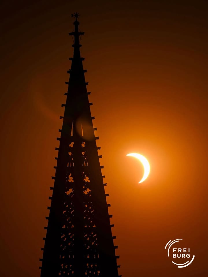 Die Silhouette eines spitzen Turms steht vor einem orangefarbenen Himmel, in dem rechts eine partielle Sonnenfinsternis als helle Sichel zu sehen ist. Unten rechts befindet sich ein weißes Logo mit dem Text "FREI BURG". (KI-Alternativtext)