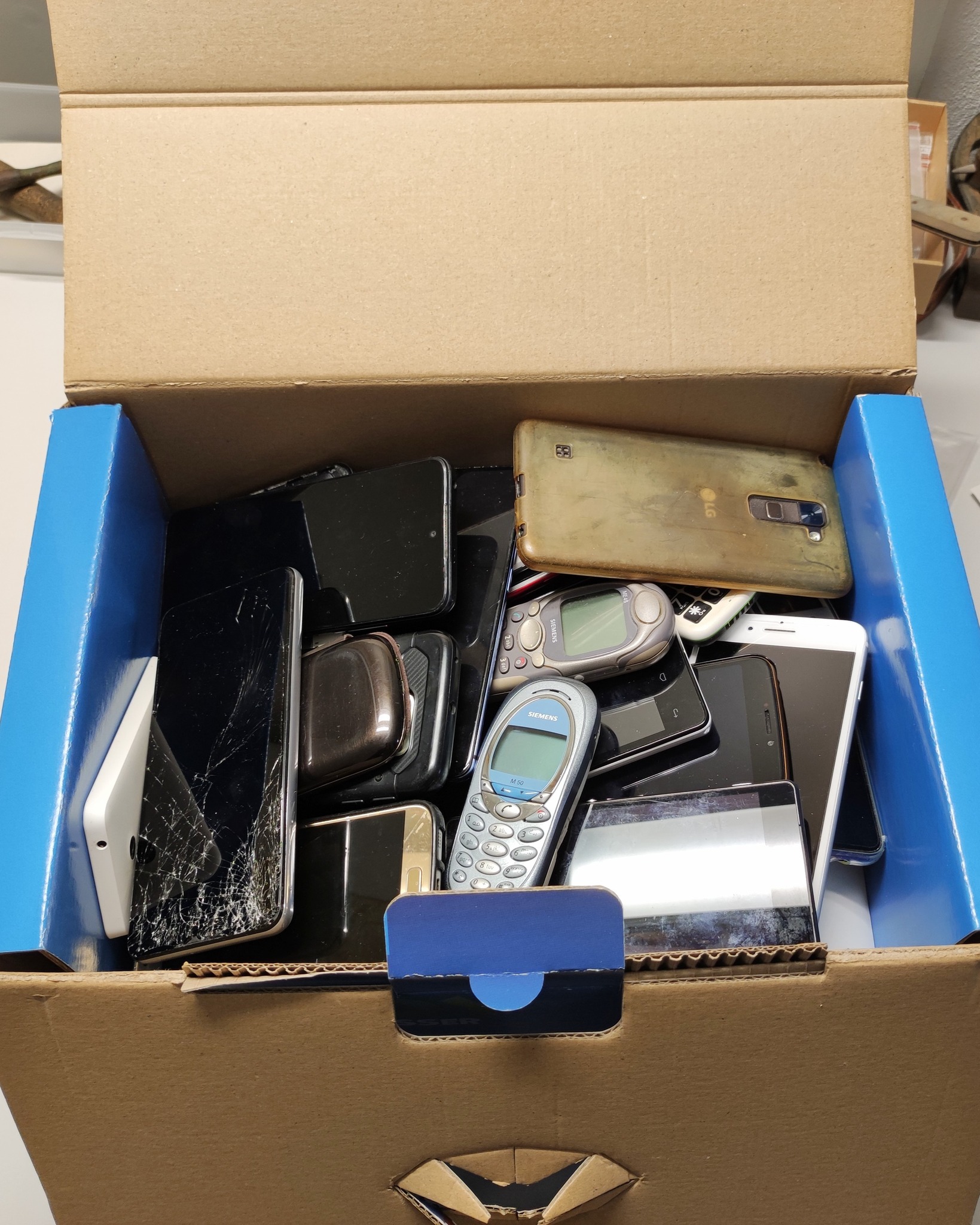 Dieser Alternativtext wurde KI-generiert: Eine Kiste, gefüllt mit verschiedenen alten Mobiltelefonen und elektronischen Geräten. Das Bild ist in Farbe gehalten.