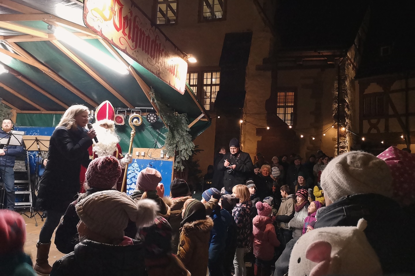 Ein Weihnachtsmann mit einem Mikrofon und einer Frau stehen vor vielen Kindern. Ein Mann im Hintergrund macht Notizen.