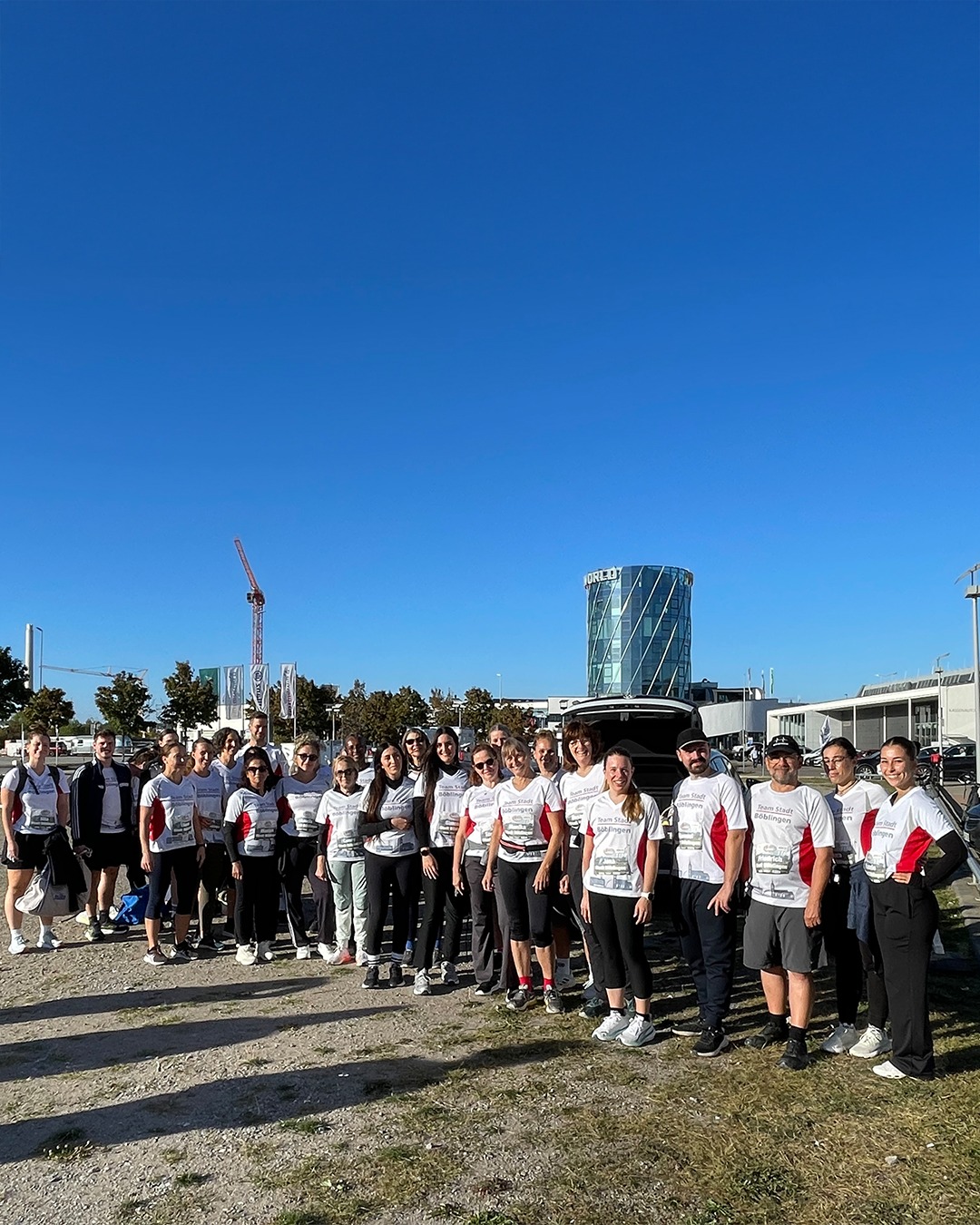 Eine Gruppe von etwa zwanzig Personen steht in mehreren Reihen im Freien auf einer Wiese bei hellem Wetter. Sie tragen weiße Sportshirts mit rotem Akzent und der Aufschrift „Team Stadt Böblingen". Im Hintergrund sind ein modernes Gebäude mit der Aufschrift „ORLETT", eine Baugerüst-Kran und Bäume unter einem blauen Himmel zu sehen. (KI-Alternativtext)
