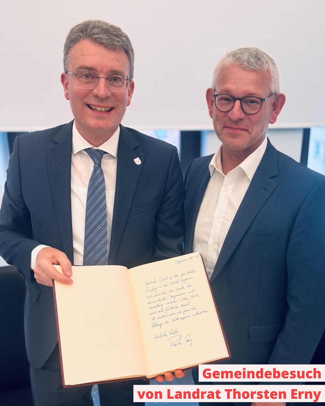 Zwei Personen in Anzügen stehen nebeneinander und halten ein offenes Buch. Es scheint sich um eine formelle Begegnung zu handeln.