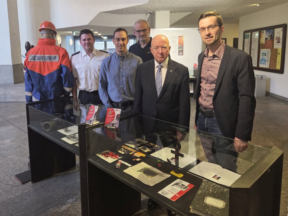 Dieser Alternativtext wurde KI-generiert: Eine Gruppe von Personen steht in einem Innenraum vor einer Vitrine mit Ausstellungsstücken. Im Hintergrund sind Bilder an der Wand zu sehen.