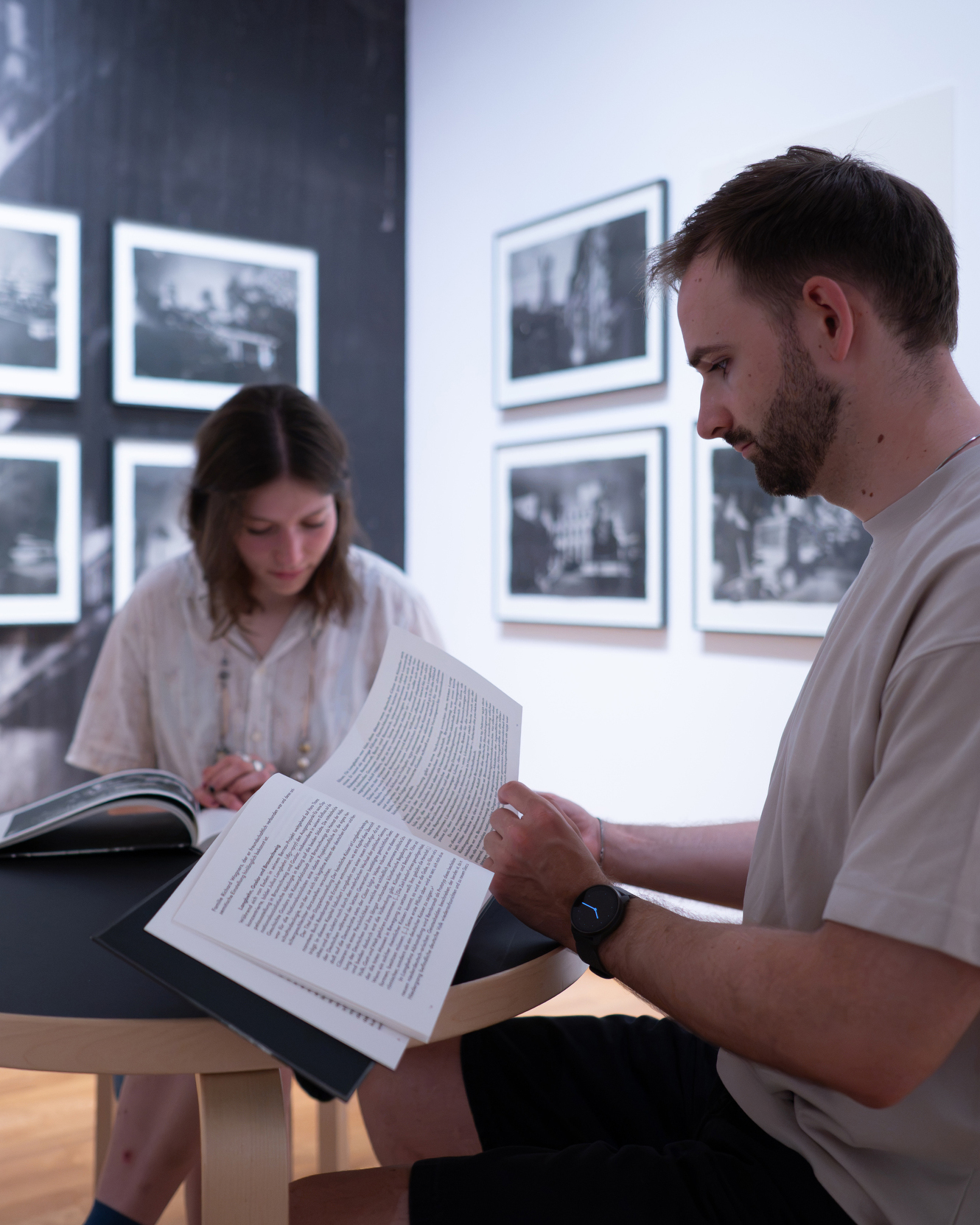 Zwei Personen sitzen an einem Tisch in einer Galerie und lesen Kataloge. Die Person im Vordergrund hält ein offenes Buch mit Textblöcken in den Händen, während im Hintergrund eine weitere Person ein Buch studiert. An den Wänden hängen mehrere gerahmte Schwarz-Weiß-Fotografien. (KI-Alternativtext)
