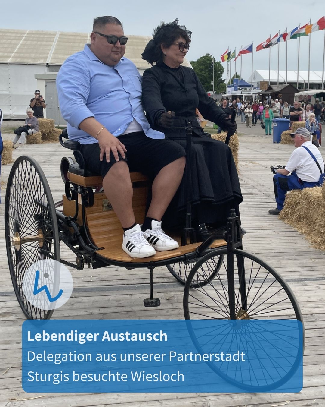 Zwei Personen sitzen in einem historischen Dreirad auf einer Holzplattform; im Hintergrund sind Menschen, Strohballen und Flaggen zu sehen. Ein blauer Textblock unten links informiert über einen lebendigen Austausch mit einer Delegation aus der Partnerstadt Sturgis in Wiesloch. (KI-Alternativtext)