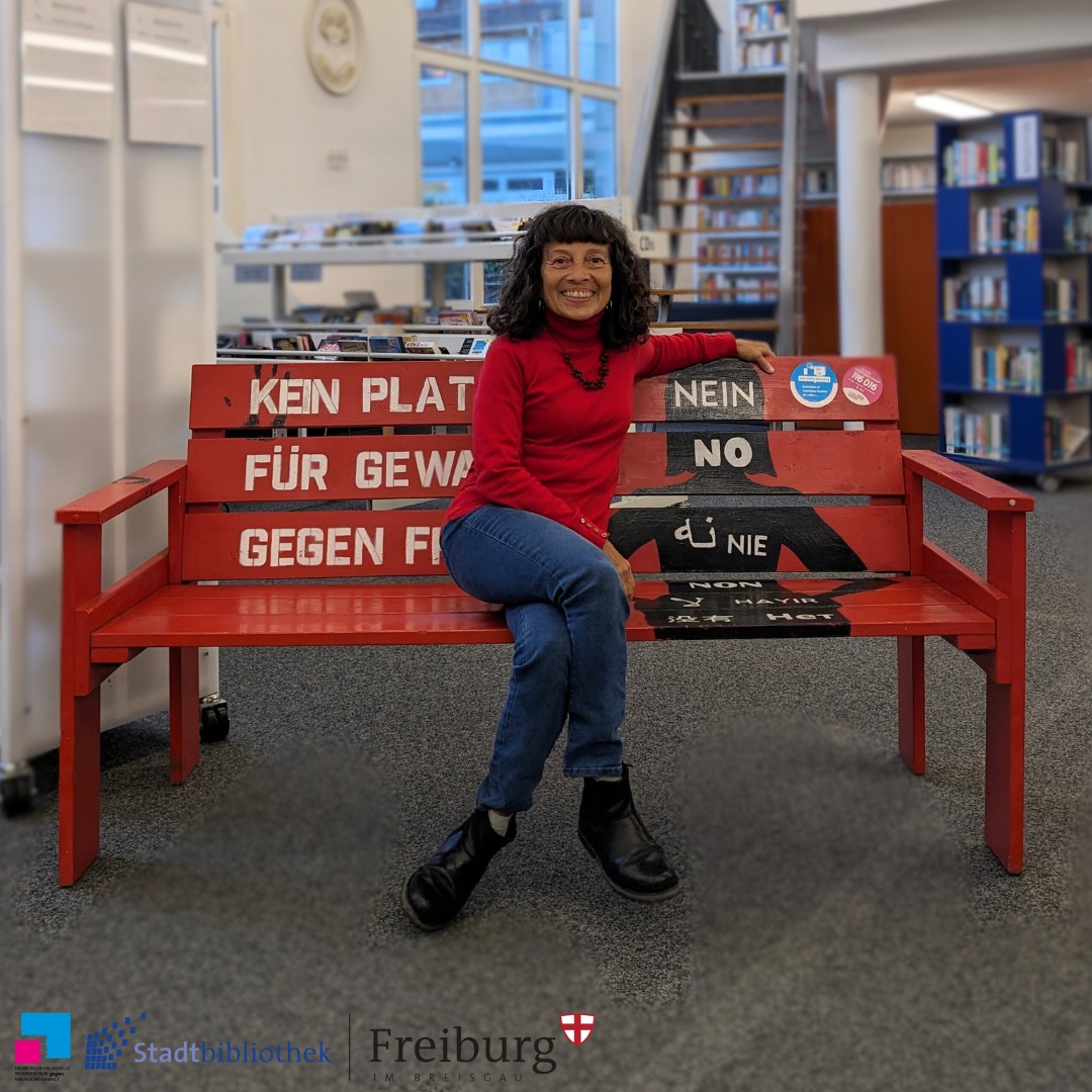 Eine Person sitzt auf einer roten Bank mit Aufschriften gegen Gewalt. Die Bank steht in einer hellen Bibliothek.