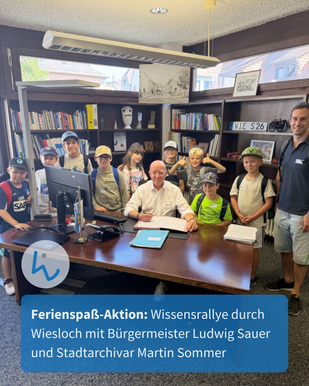 Eine Person unterzeichnet an einem Schreibtisch in einem Raum mit Bücherregalen ein Dokument, während eine Gruppe von Kindern und eine weitere Person daneben stehen. Ein blauer Textbalken unten lautet: "Ferienspaß-Aktion: Wissensrallye durch Wiesloch mit Bürgermeister Ludwig Sauer und Stadtarchivar Martin Sommer". (KI-Alternativtext)