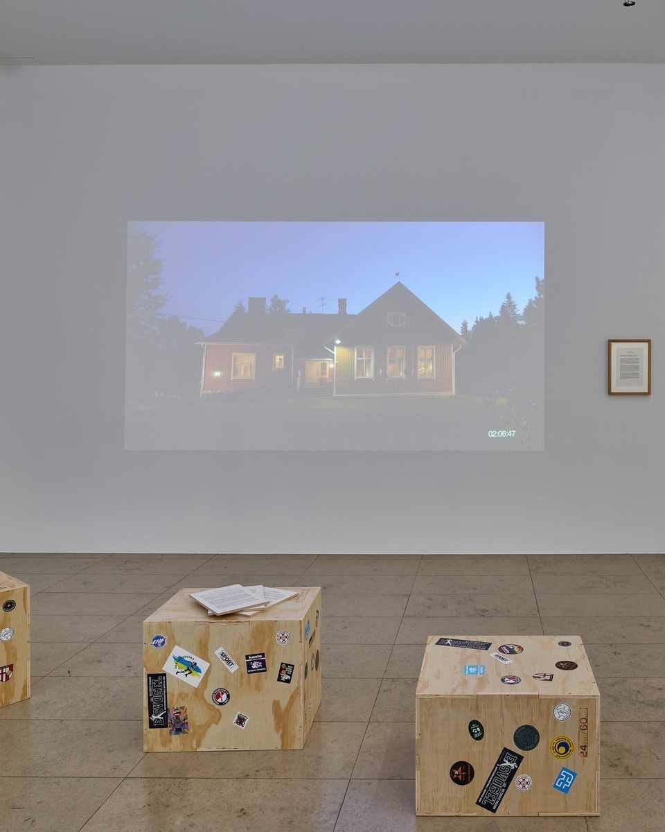 Eine große Wandprojektion zeigt ein Haus bei Dämmerung mit leuchtenden Fenstern und der Zeitangabe 02:06:47 in der unteren rechten Ecke. Rechts daneben hängt ein gerahmter Text an der weißen Wand. Im Vordergrund stehen drei mit verschiedenen Aufklebern beklebte Holzkisten auf dem Fliesenboden, auf der mittleren Kiste liegen lose Papiere. (KI-Alternativtext)