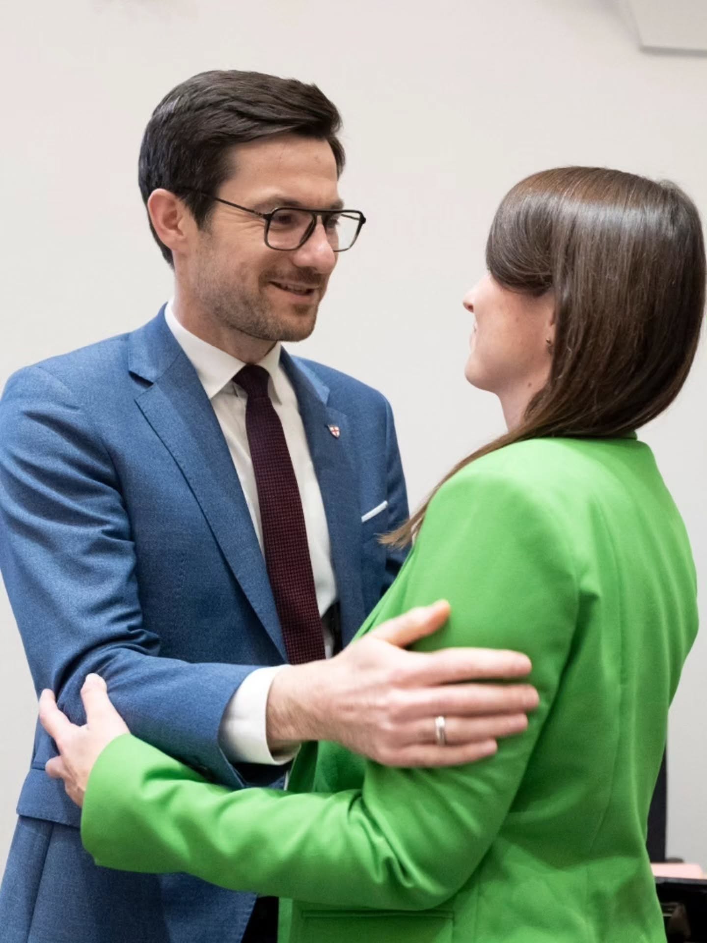 Dieser Alternativtext wurde KI-generiert: Zwei Personen in formeller Kleidung unterhalten sich freundlich. Eine Person umarmt die andere leicht.