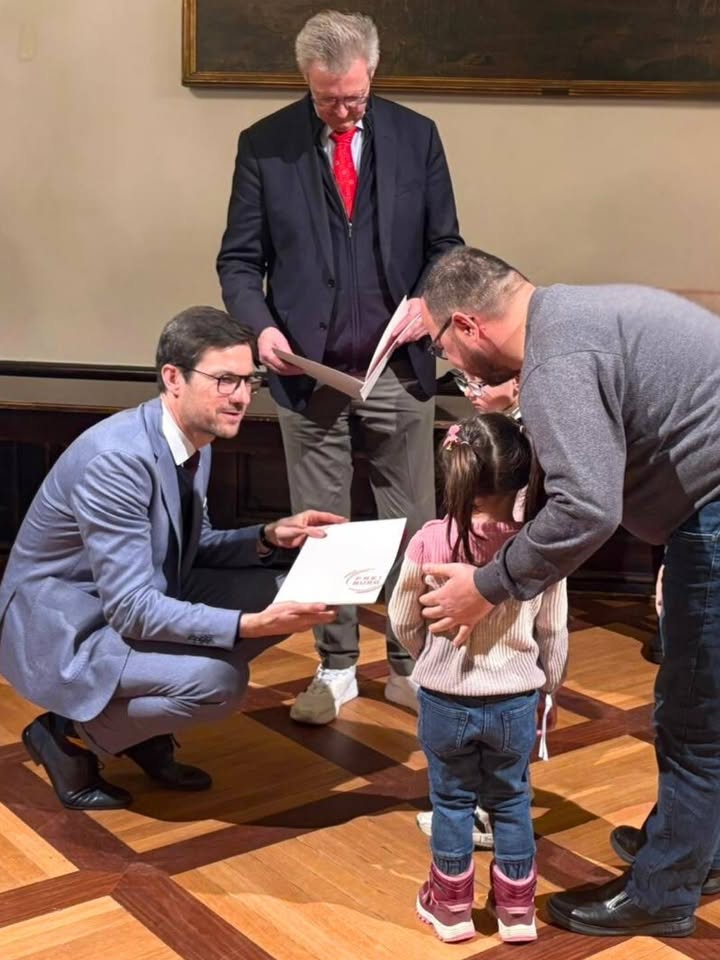 Dieser Alternativtext wurde KI-generiert: Mehrere Personen stehen und knien in einem Raum mit Parkettboden. Eine Person erhält ein Dokument, während eine andere eine kleine Person umarmt.