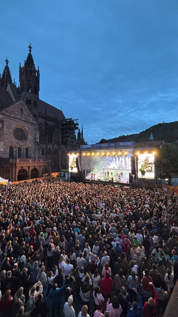 Große Menschenmenge vor einer beleuchteten Bühne bei Dämmerung, dahinter gotische Kirche mit Turm. Auf der Bühne sind Musiker und große Bildschirme mit Live-Übertragung zu sehen. (KI-Alternativtext)
