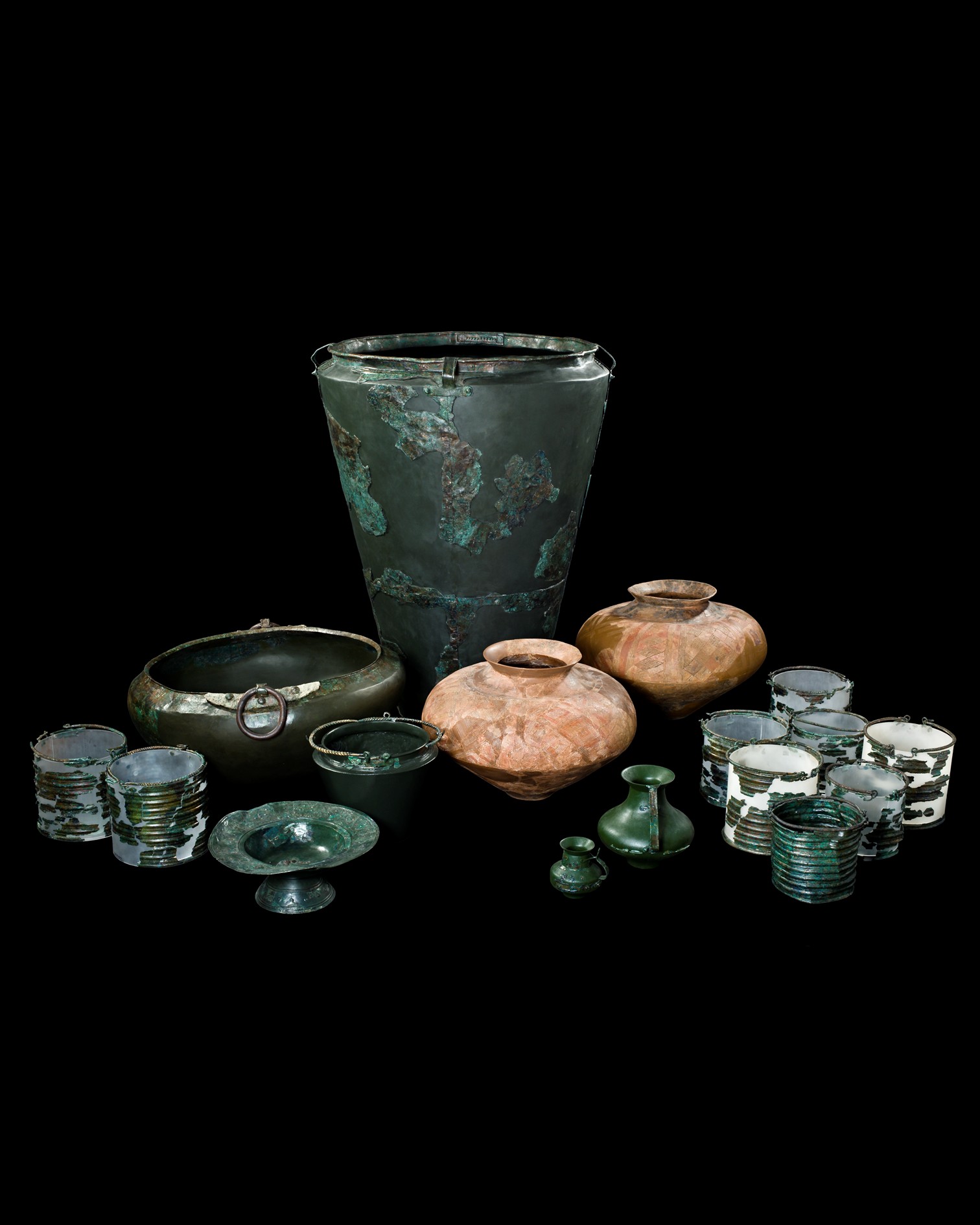 Dieser Alternativtext wurde KI-generiert: Schwarz-weißes Stillleben mit verschiedenen alten Gefäßen und Bechern aus Metall und Keramik. Die Objekte sind auf einer dunklen Oberfläche angeordnet.