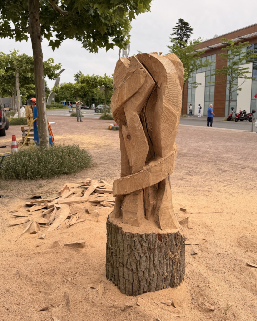 Holzskulptur auf Baumstumpf, abstrakt geschnitzt mit sichtbaren Spänen am Boden, im Hintergrund Menschen und modernes Gebäude. (KI-Alternativtext)