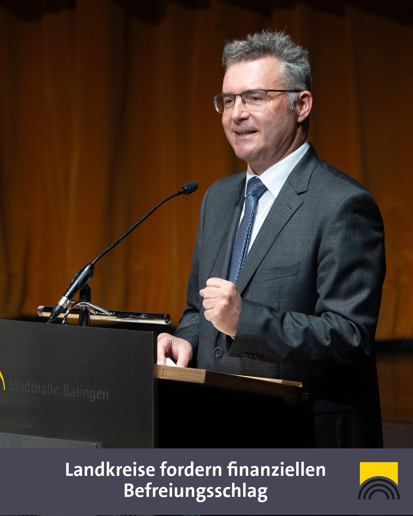 Dieser Alternativtext wurde KI-generiert: Eine Person mit Brille hält eine Rede am Rednerpult. Im Hintergrund ist ein goldfarbener Vorhang zu sehen.