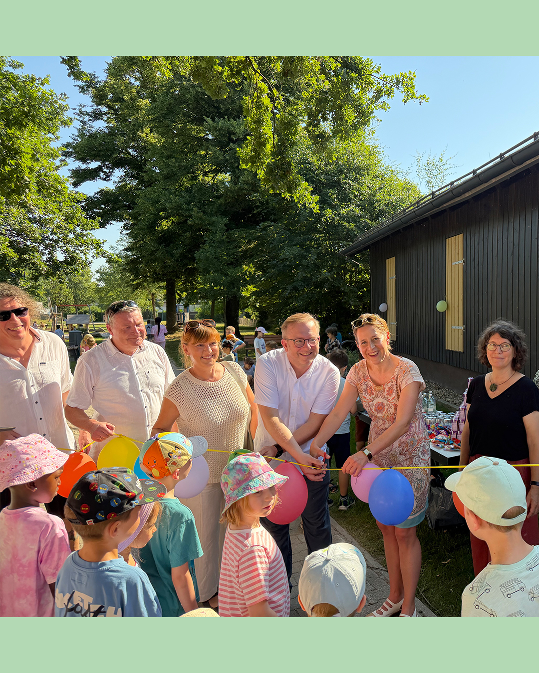 Mehrere Erwachsene stehen im Freien vor einem Gebäude und halten gemeinsam ein gelbes Band mit bunten Luftballons. Davor stehen mehrere Kinder mit Kopfbedeckungen und blicken zur Gruppe, was auf eine Feierlichkeit hindeutet. (KI-Alternativtext)