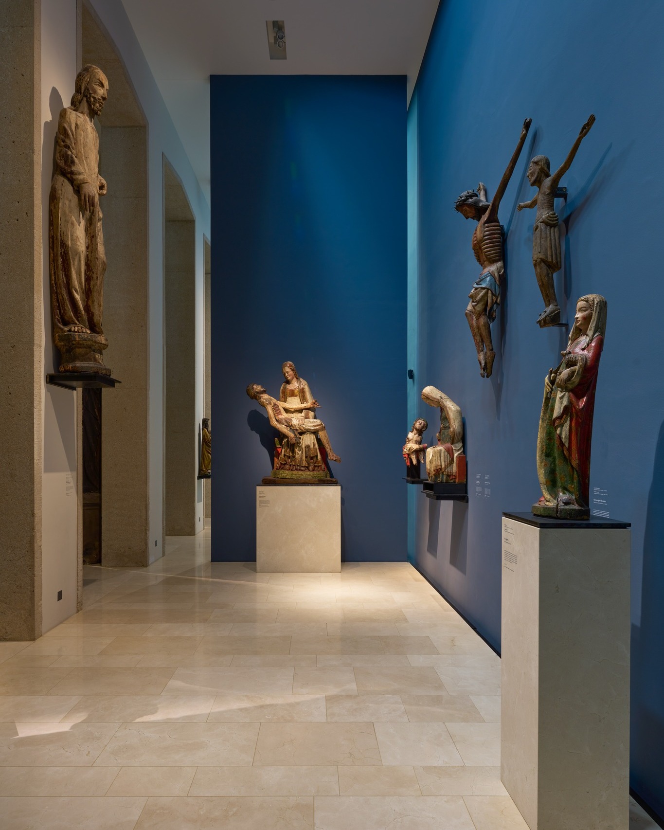 Dieser Alternativtext wurde KI-generiert: Im Innenraum eines Museums stehen mehrere alte Skulpturen auf Sockeln. Ein blauer Akzentwall bildet den Hintergrund der Ausstellung.